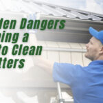 A man in blue work clothes climbs a ladder to clean house gutters, highlighting the ladder fall risk associated with gutter cleaning. Green text reads: The Hidden Dangers of Climbing a Ladder to Clean Your Gutters. K-Guard logo is at the bottom left.